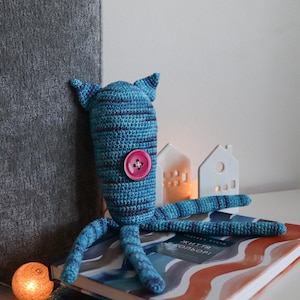 Peut inclure: Une pieuvre au crochet bleue et violette faite à la main, avec un bouton rose. La pieuvre a un motif rayé et est posée sur un livre. Des maisons blanches décoratives et un globe lumineux sont en arrière-plan.