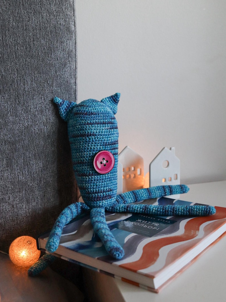 May include: A handmade blue and purple crochet octopus toy with a pink button. The octopus has a striped pattern and is sitting on a book. Decorative white houses and a glowing orb are in the background.