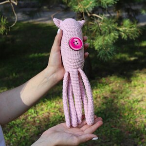 May include: A handmade, light pink crocheted octopus toy with a large pink button detail. The octopus has eight long, dangling tentacles and small, pointed ears. The toy is held in a person's hands against a blurred green background.