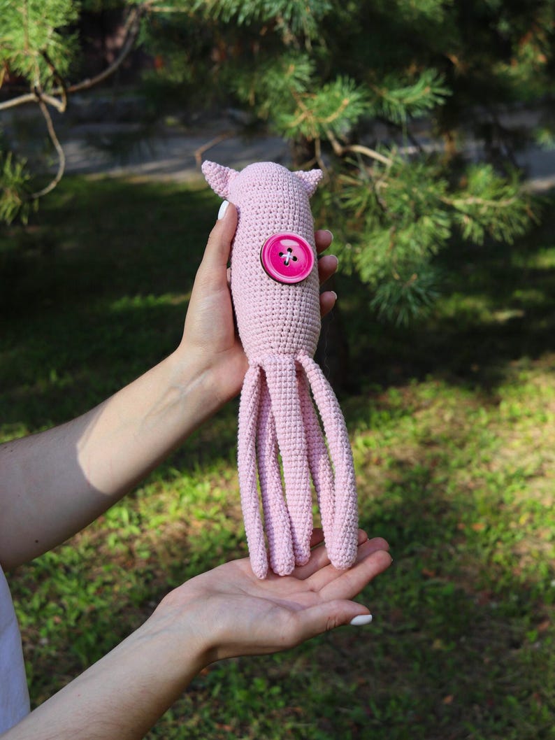 May include: A handmade, light pink crocheted octopus toy with a large pink button detail. The octopus has eight long, dangling tentacles and small, pointed ears. The toy is held in a person's hands against a blurred green background.