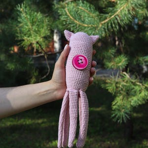 May include: A handmade, light pink crocheted octopus toy with a large pink button detail. The octopus has a rounded head with small ears and long, dangling tentacles. The toy is held against a blurred green background of trees.