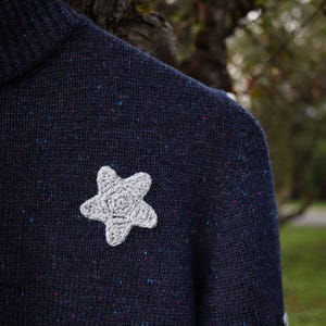 May include: A navy blue sweater with a gray crocheted star applique. The sweater has a speckled pattern with flecks of blue, pink, and other colors. The star is centered on the chest of the sweater.