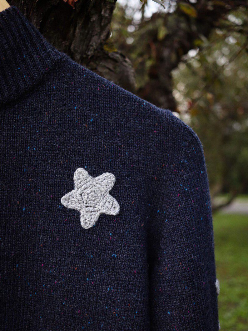 May include: A navy blue sweater with a gray crocheted star applique. The sweater has a speckled pattern with flecks of blue, pink, and other colors. The star is centered on the chest of the sweater.