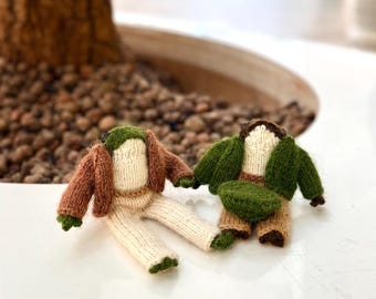 May include: Two handmade, miniature knitted dolls. One doll wears a brown jacket, green hat, and cream-colored pants. The other doll wears a green jacket, brown pants, and a green hat. The dolls are resting on a white surface.