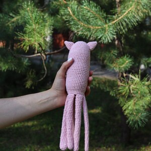May include: A handmade, light pink crochet octopus toy with eight long tentacles and small ear-like protrusions. The soft toy is held against a backdrop of green foliage and sunlight.