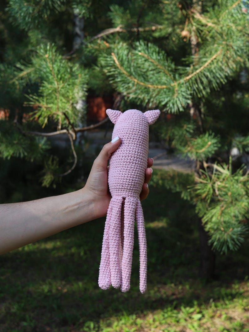 May include: A handmade, light pink crochet octopus toy with eight long tentacles and small ear-like protrusions. The soft toy is held against a backdrop of green foliage and sunlight.