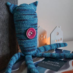 May include: A handmade blue and purple crochet monster doll with a large pink button. The doll has two pointed ears and multiple tentacles. The doll is sitting on a book with text in a foreign language.