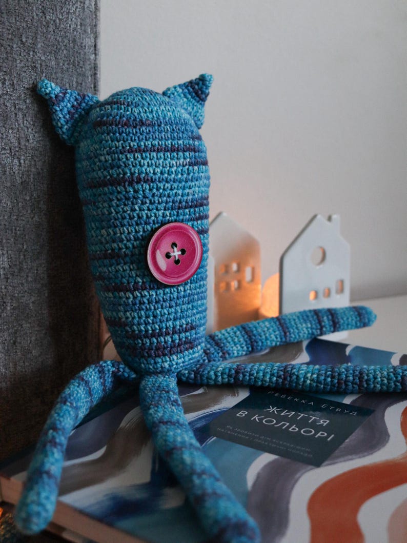 May include: A handmade blue and purple crochet monster doll with a large pink button. The doll has two pointed ears and multiple tentacles. The doll is sitting on a book with text in a foreign language.