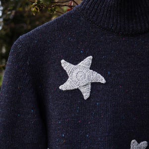 May include: Navy blue sweater with a high neck and three gray crocheted stars. The sweater has a speckled pattern with flecks of blue, red, and yellow. The stars are textured and add a whimsical touch to the garment.