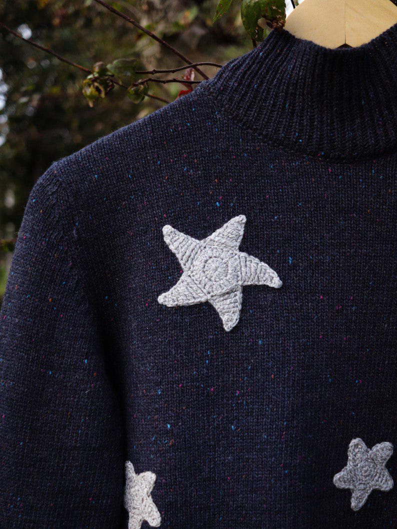 May include: Navy blue sweater with a high neck and three gray crocheted stars. The sweater has a speckled pattern with flecks of blue, red, and yellow. The stars are textured and add a whimsical touch to the garment.