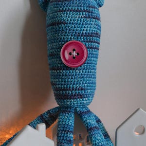 May include: A handmade blue and purple striped crochet creature with a large pink button eye and small pointed ears. The creature has multiple tentacles and is positioned in front of two white ceramic house-shaped candle holders.