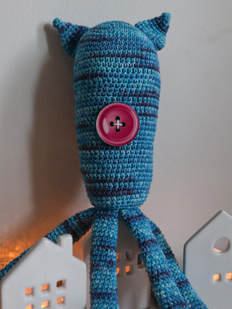 May include: A handmade blue and purple striped crochet creature with a large pink button eye and small pointed ears. The creature has multiple tentacles and is positioned in front of two white ceramic house-shaped candle holders.