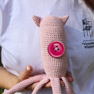 May include: A handmade, light pink crocheted octopus plush toy with a large, round, hot pink button. The octopus has small ears and tentacles. The toy is held against a white t-shirt.