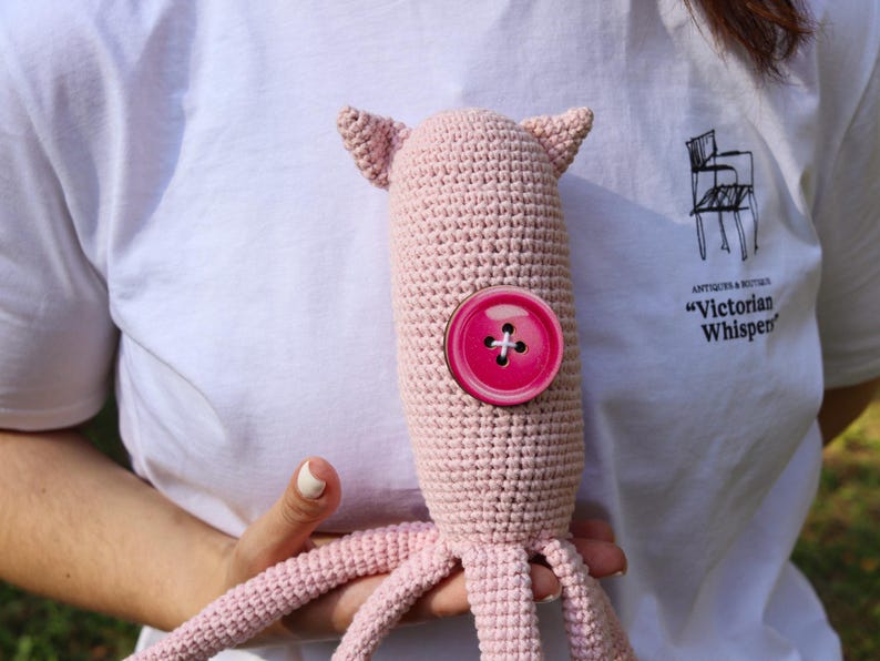 May include: A handmade, light pink crocheted octopus plush toy with a large, round, hot pink button. The octopus has small ears and tentacles. The toy is held against a white t-shirt.