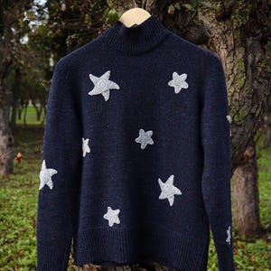 May include: A navy blue sweater with a turtleneck collar and white star appliqu&eacute;s. The sweater is hanging on a wooden hanger against a blurred background of trees and foliage. The stars are of varying sizes.