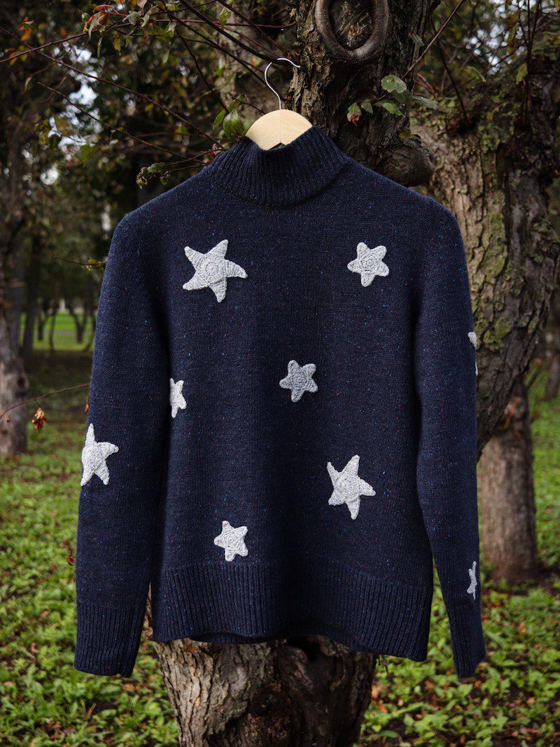 May include: A navy blue sweater with a turtleneck collar and white star appliqu&eacute;s. The sweater is hanging on a wooden hanger against a blurred background of trees and foliage. The stars are of varying sizes.