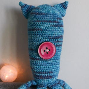 May include: A handmade blue and purple striped crochet creature with a pink button eye. The whimsical design features two small ears and multiple tentacle-like appendages. A soft, glowing orb is in the background.