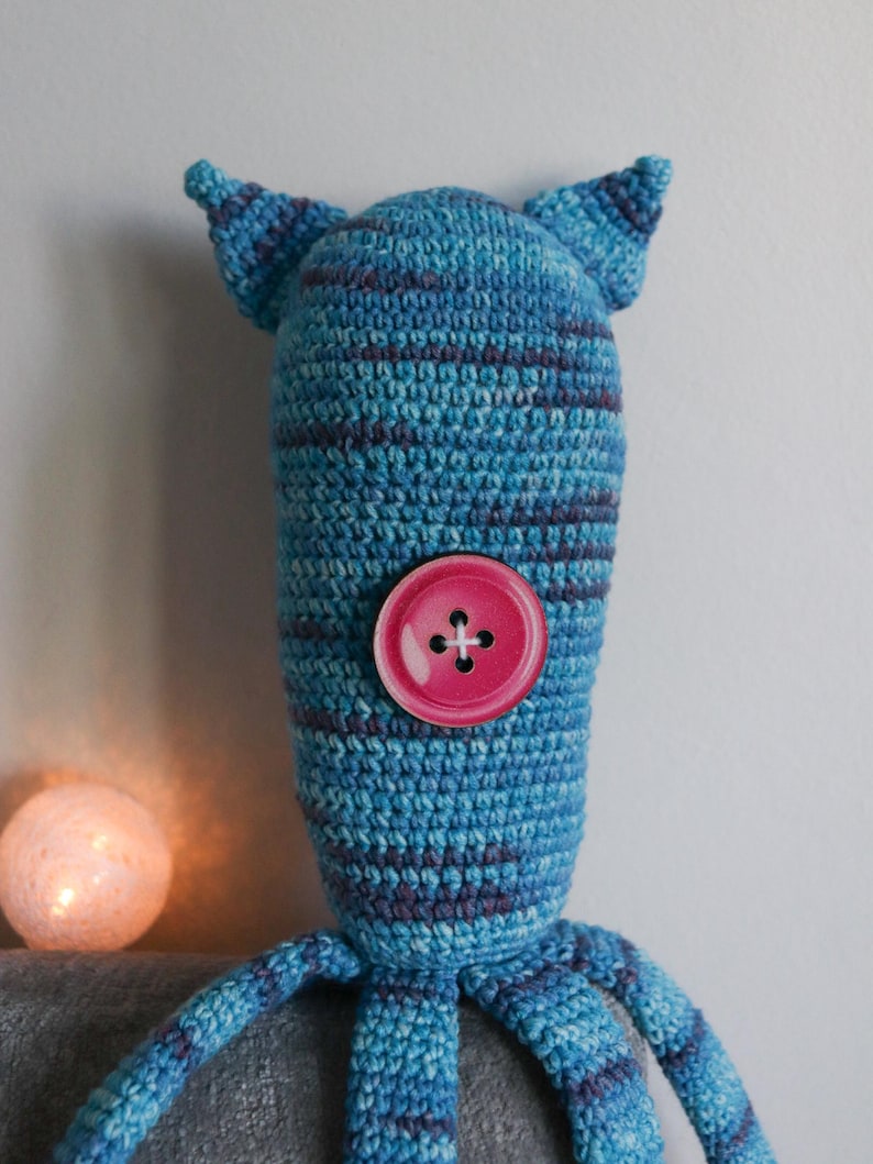 May include: A handmade blue and purple striped crochet creature with a pink button eye. The whimsical design features two small ears and multiple tentacle-like appendages. A soft, glowing orb is in the background.