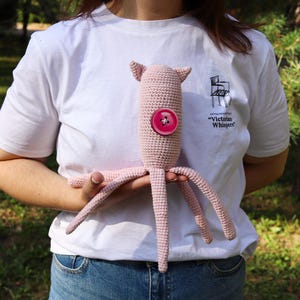 May include: A handmade pink crochet octopus plush toy with a large pink button on its body. The toy has four tentacles and two small ears. The person holding the toy is wearing a white t-shirt with the text "Victorian Whispers".