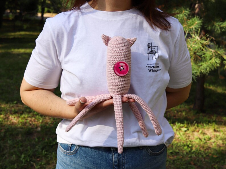 May include: A handmade pink crochet octopus plush toy with a large pink button on its body. The toy has four tentacles and two small ears. The person holding the toy is wearing a white t-shirt with the text "Victorian Whispers".