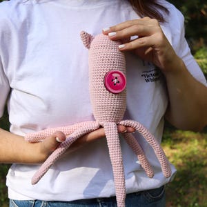 May include: A handmade, light pink crochet octopus toy with a large, round, pink button. The octopus has a cylindrical body, small ear-like protrusions, and multiple tentacles. The toy is held against a background of green foliage.
