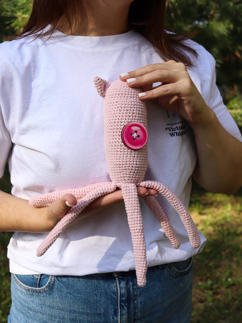 May include: A handmade, light pink crochet octopus toy with a large, round, pink button. The octopus has a cylindrical body, small ear-like protrusions, and multiple tentacles. The toy is held against a background of green foliage.