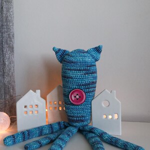 May include: A handmade blue and navy crochet octopus plush toy with a pink button. The octopus has a striped pattern on its head and tentacles. Decorative white ceramic houses with lit windows are in the background.