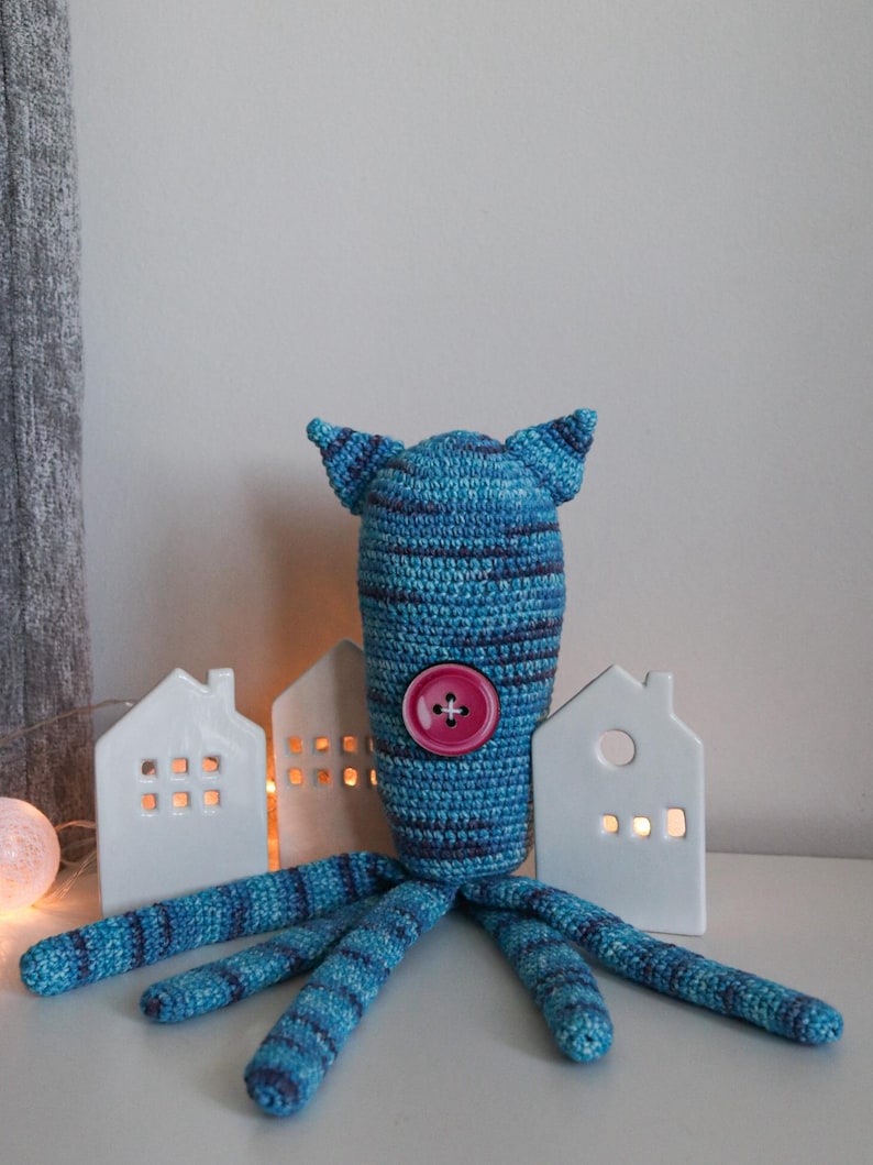 May include: A handmade blue and navy crochet octopus plush toy with a pink button. The octopus has a striped pattern on its head and tentacles. Decorative white ceramic houses with lit windows are in the background.