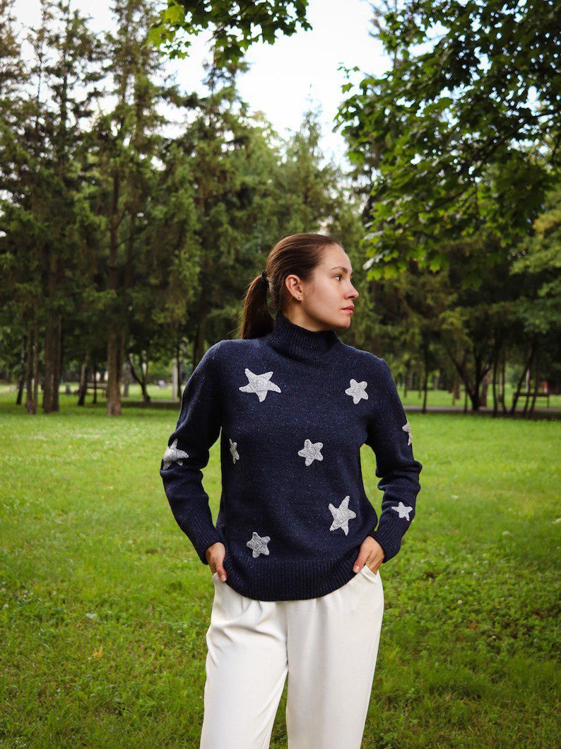 May include: A navy blue turtleneck sweater with white star patterns. The sweater is paired with white pants. The image is taken outdoors with a green grassy background and trees.