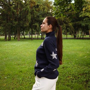 May include: A navy blue sweater with white star accents is paired with white pants. The sweater has a turtleneck and the star designs are on the sleeves and chest. The person is standing in a grassy park.