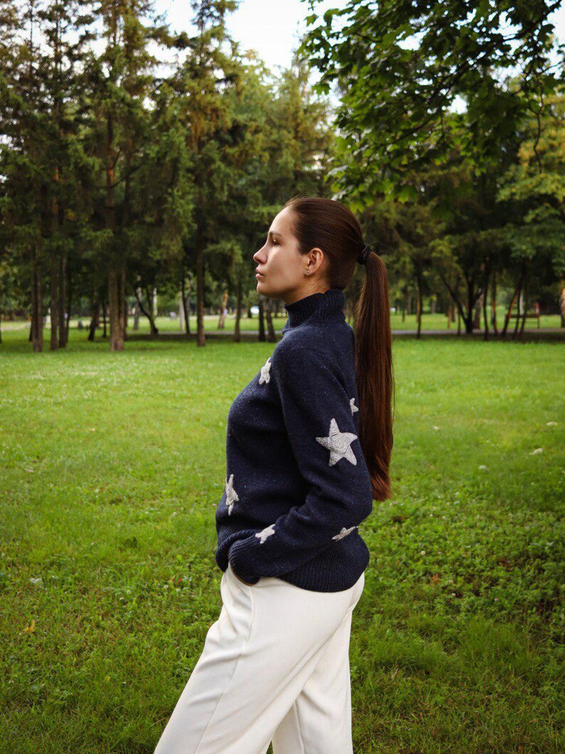 May include: A navy blue sweater with white star accents is paired with white pants. The sweater has a turtleneck and the star designs are on the sleeves and chest. The person is standing in a grassy park.