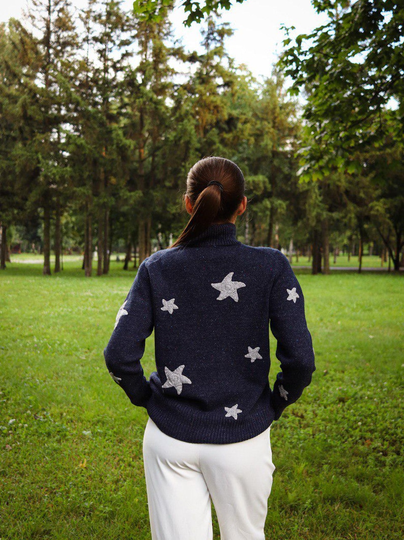 May include: A navy blue sweater with a turtleneck and white star designs. The person is wearing white pants and standing in a grassy park with trees in the background. The sweater has white stars on the sleeves and back.