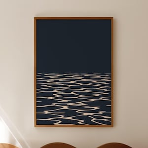 May include: A framed art print featuring a minimalist abstract design. The artwork depicts a dark blue expanse above a wavy, light beige pattern, resembling water. The wooden frame adds a natural touch to the piece, suitable for home decor.