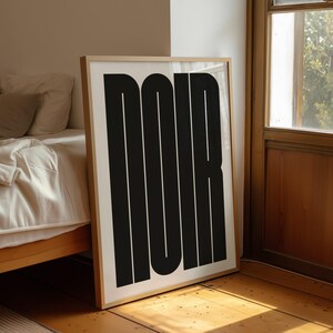 May include: A framed art print featuring the word "NOIR" in bold, black, vertical block letters against a white background. The artwork is displayed on a wooden floor, leaning against a wall near a bed and a window.