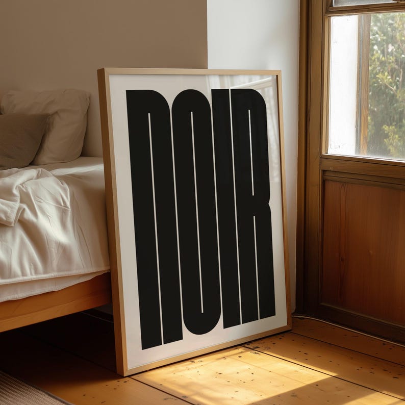 May include: A framed art print featuring the word "NOIR" in bold, black, vertical block letters against a white background. The artwork is displayed on a wooden floor, leaning against a wall near a bed and a window.
