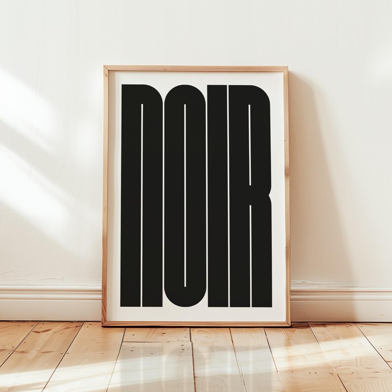 May include: A framed art print featuring the word "NOIR" in bold, black, sans-serif letters. The artwork is displayed against a white background and framed in a light-colored wood frame. The print is leaning against a white wall on a wooden floor.