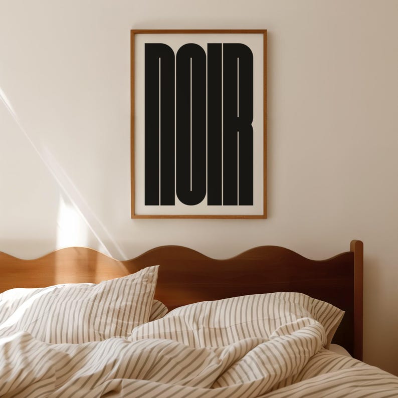 May include: A framed art print with the word "NOIR" in bold, black, sans-serif letters. The artwork is displayed on a light-colored wall above a bed with striped bedding and a wooden headboard. The frame is a light brown color.