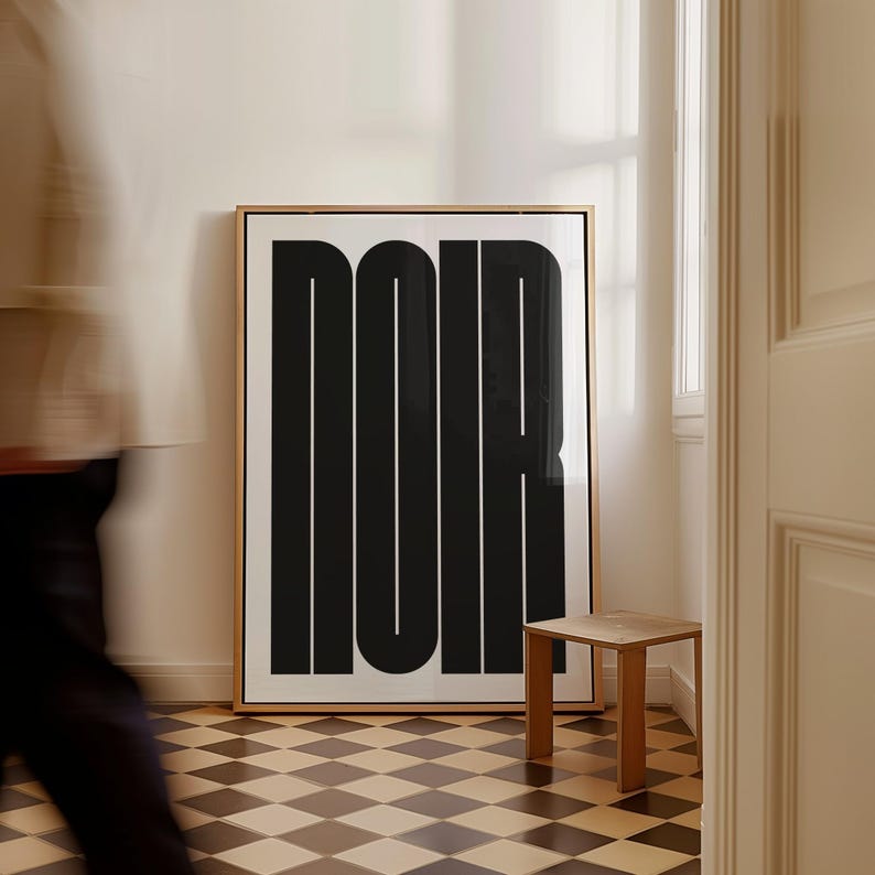 May include: A framed art print with the word "NOIR" in bold, black letters. The artwork is displayed in a room with a checkered floor and a small wooden stool. The print has a gold frame and is leaning against a white wall.