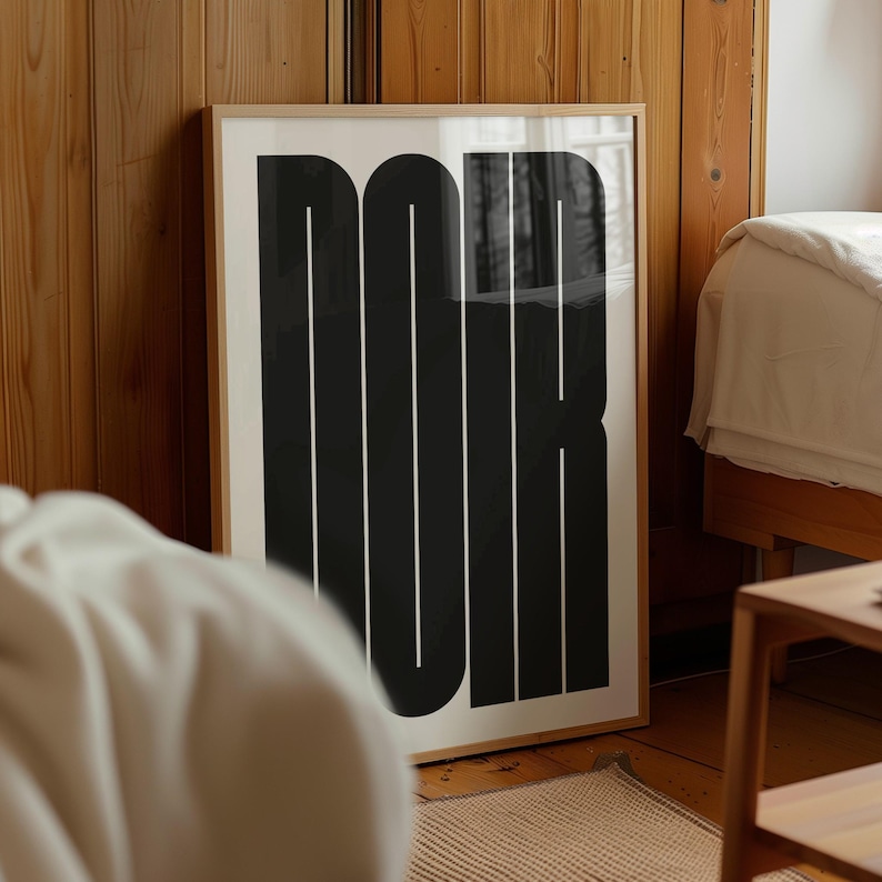 May include: A framed art print with the word "NOIR" in bold, black, sans-serif letters. The letters are set against a white background, with a thin white line running through each letter. The frame is a light wood color.
