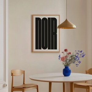 May include: A framed art print with the word "NOIR" in bold black letters hangs on a beige wall. A round white table with wooden legs is set with a blue vase of flowers. A gold pendant light hangs above.