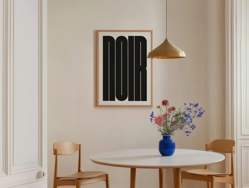 May include: A framed art print with the word "NOIR" in bold black letters hangs on a beige wall. A round white table with wooden legs is set with a blue vase of flowers. A gold pendant light hangs above.