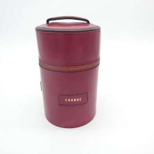 Op de afbeelding: Een cilindervormige bordeauxrode koker met een handvat aan de bovenkant en een ritssluiting. De koker heeft een rechthoekig label aan de voorzijde met het woord COGNAC in gouden letters. Het ontwerp bevat donkere biezen langs de randen.