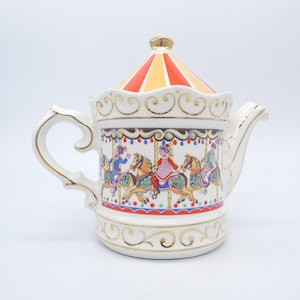 May include: A white ceramic teapot with a carousel design. The lid is striped red and yellow, and the body features a carousel scene with riders on horses. Gold accents adorn the handle, spout, and base.
