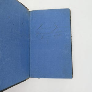 K&ouml;nnte beinhalten: Offenes antikes Buch mit blauen Seiten und handschriftlichem Text. Der Einband des Buches ist dunkel, und die Seiten weisen Alterserscheinungen auf. Der Text ist in Kursivschrift gehalten und unterstreicht die Vintage-&Auml;sthetik. Das Buch ist ge&ouml;ffnet und gibt den Blick ins Innere frei.