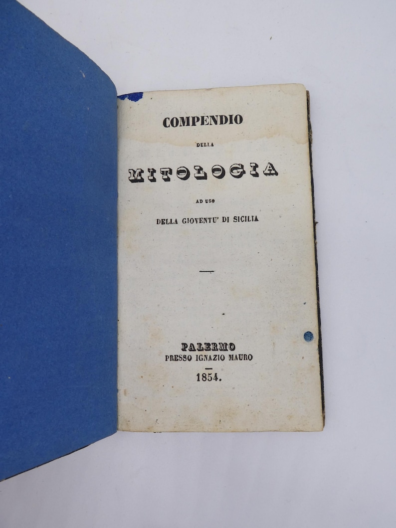 K&ouml;nnte beinhalten: Ein aufgeschlagenes Buch mit blauem Einband und gealterten Seiten. Die Titelseite tr&auml;gt die Aufschrift "COMPENDIO DELLA MITOLOGIA" mit dem Text "AD USO DELLA GIOVENTU' DI SICILIA" und "PALERMO PRESSO IGNAZIO MAURO 1854."