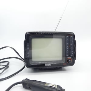 Könnte beinhalten: Ein schwarzer tragbarer Fernseher mit einer silbernen Antenne und dem Markennamen "IRRADIO" auf der Vorderseite. Der Fernseher hat einen Bildschirm, eine Lautstärkeregelung und ein Netzkabel mit einem Auto-Adapter.