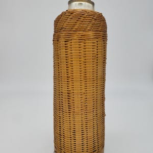 Könnte beinhalten: Eine hohe, zylindrische Flasche mit einem gewebten Korbgeflecht-Äußeren. Die Flasche hat einen Holzsockel und einen Korken mit einer silberfarbenen Metallkappe. Das Korbgeflecht ist hellbraun.