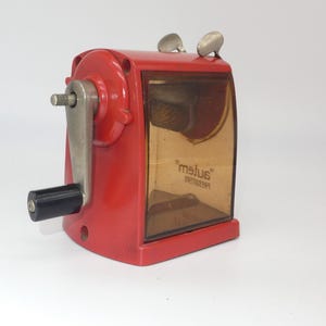 Peut inclure: Un taille-crayon en métal rouge avec une fenêtre en plastique transparent et une poignée noire. Le taille-crayon a une manivelle sur le côté et le texte "autem" imprimé sur le devant.