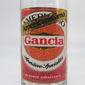 May include: A glass of Gancia Americanissimo Aperitivo Specialita, a red and gold label with the text "Americanissimo" and "Gancia" in black. The label also includes the text "Aperitivo Specialita" and "Specialita dell'Antica Casa Gancia & C. Savas Spa - Canelli (Italia)".