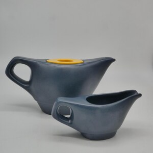 Può includere: Due brocche di ceramica blu. La brocca più grande ha un coperchio giallo. Entrambe le brocche hanno un manico e un beccuccio. Le brocche hanno una finitura opaca e un design semplice e moderno.